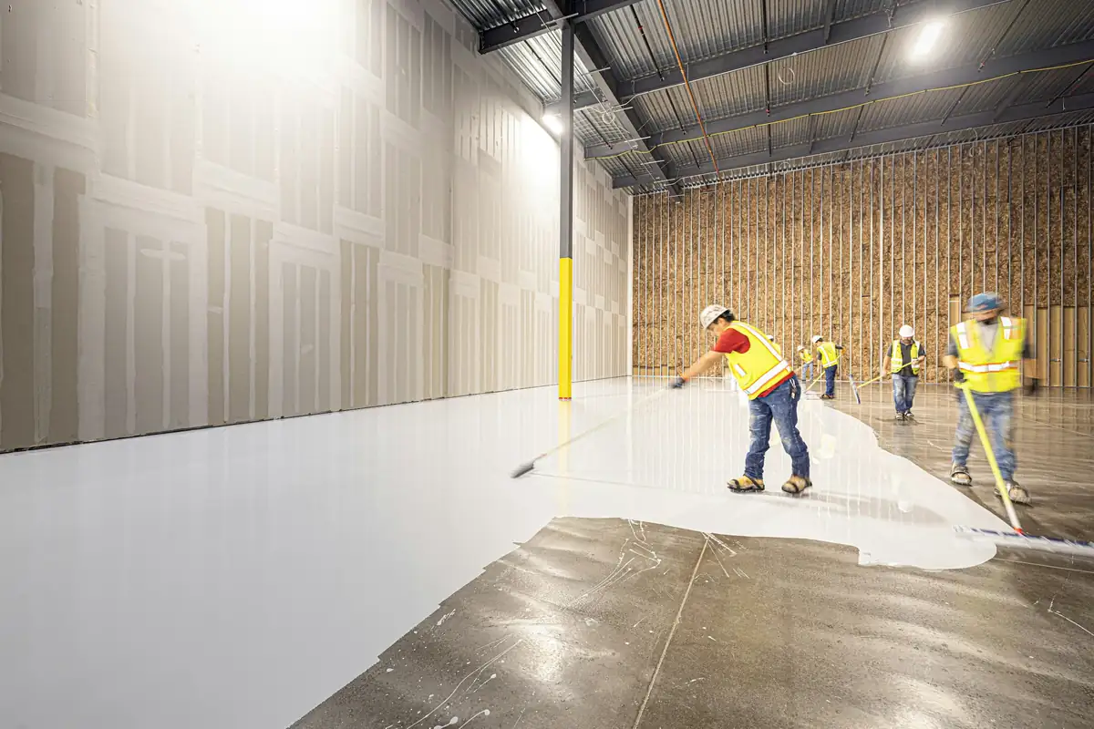 Workers spreading white floor coating over a bare concrete slab with squeegees and rollers