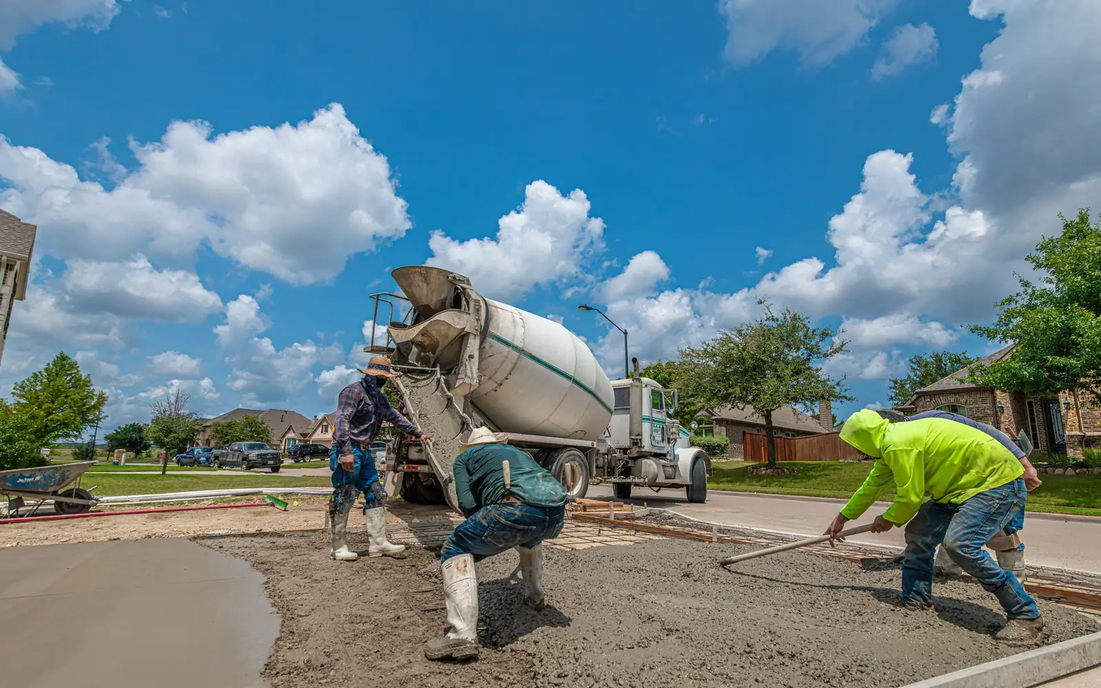 Crew in rubber boots spreading wet concrete from a mixer truck chute into a formed residential driveway