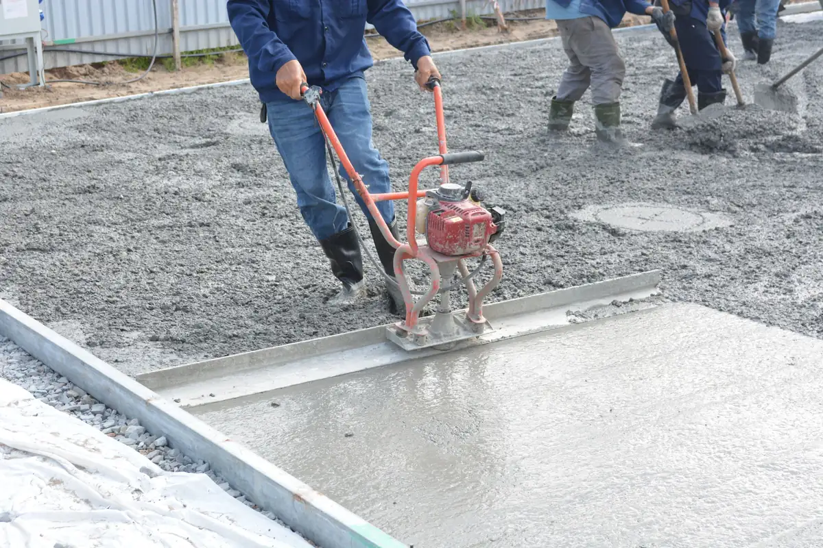 Worker running a gas-powered vibrating screed across a freshly poured slab between steel forms